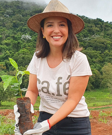 Coffee Community farmer Carolina Alckmin wearing a straw hat and holding a young coffee plant at Santo Antonio farm.