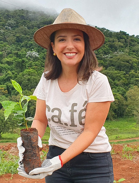 Coffee Community farmer Carolina Alckmin wearing a straw hat and holding a young coffee plant at Santo Antonio farm.
